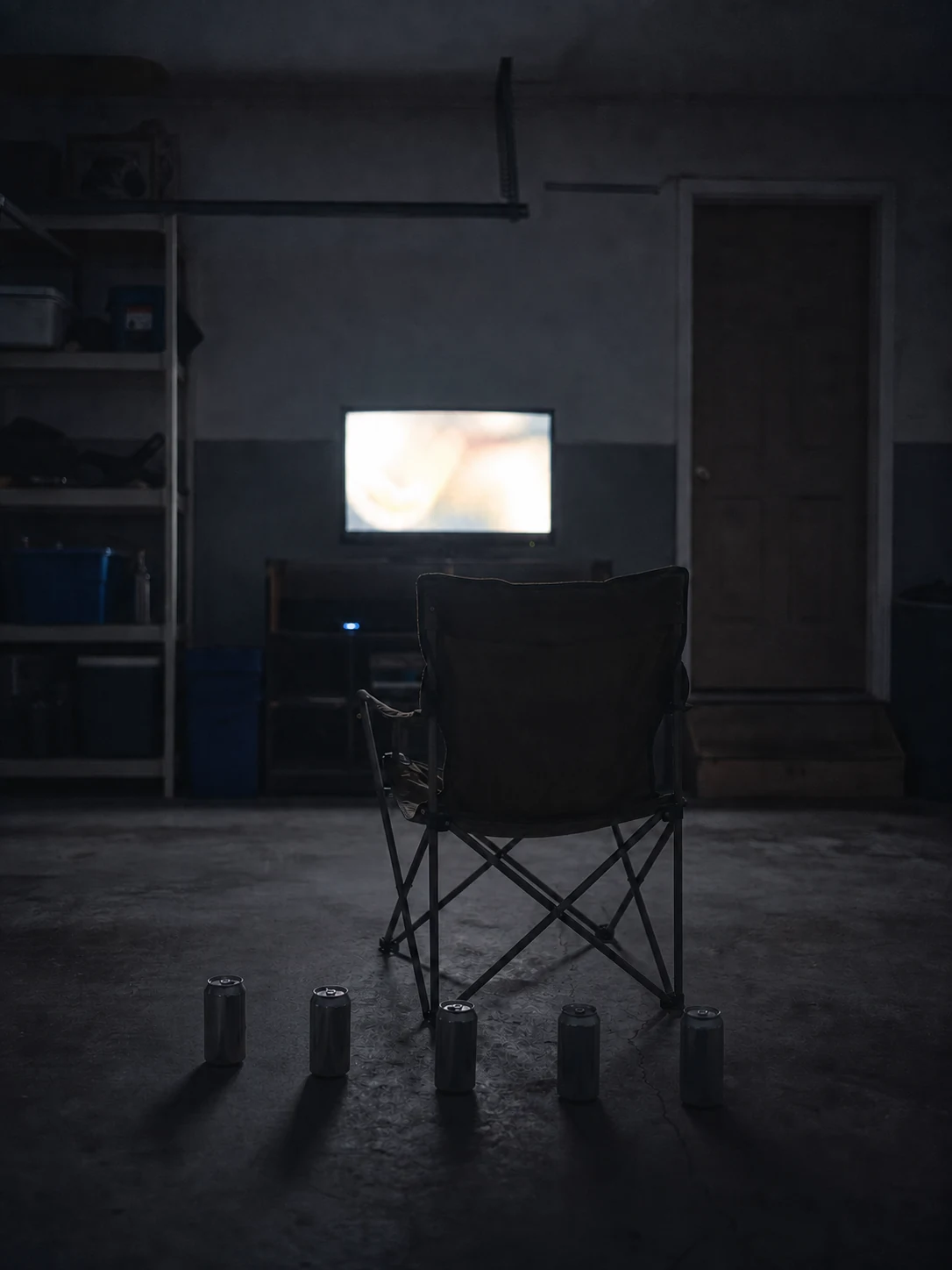 An empty garage chair faces a softly glowing television, with five unbranded cans lined on the concrete floor.